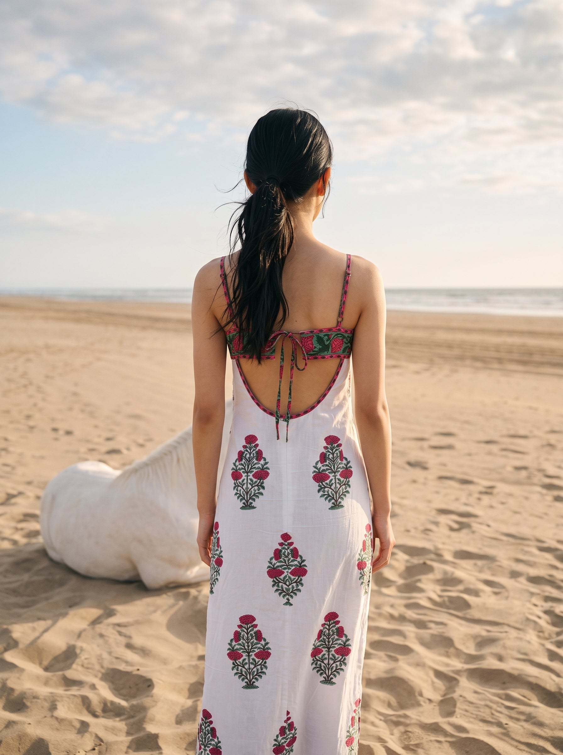 A woman stands on a sandy beach facing the ocean, wearing a white maxi dress with red and green floral embroidery and a red embroidered halter-style back detail.