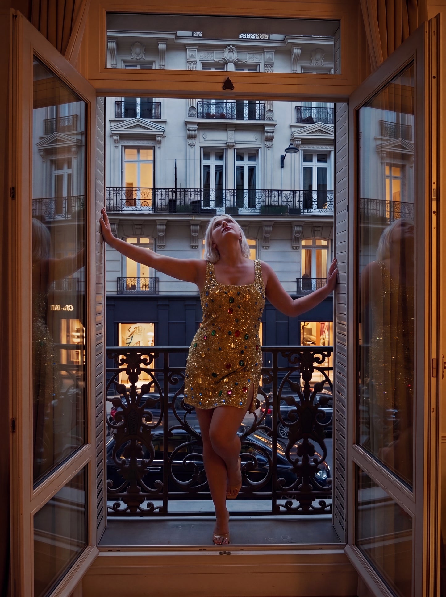 A woman in a sparkling gold sequined mini dress poses on a balcony with open French doors, looking upward with her arms outstretched. The background shows a classic Parisian-style building facade at dusk.