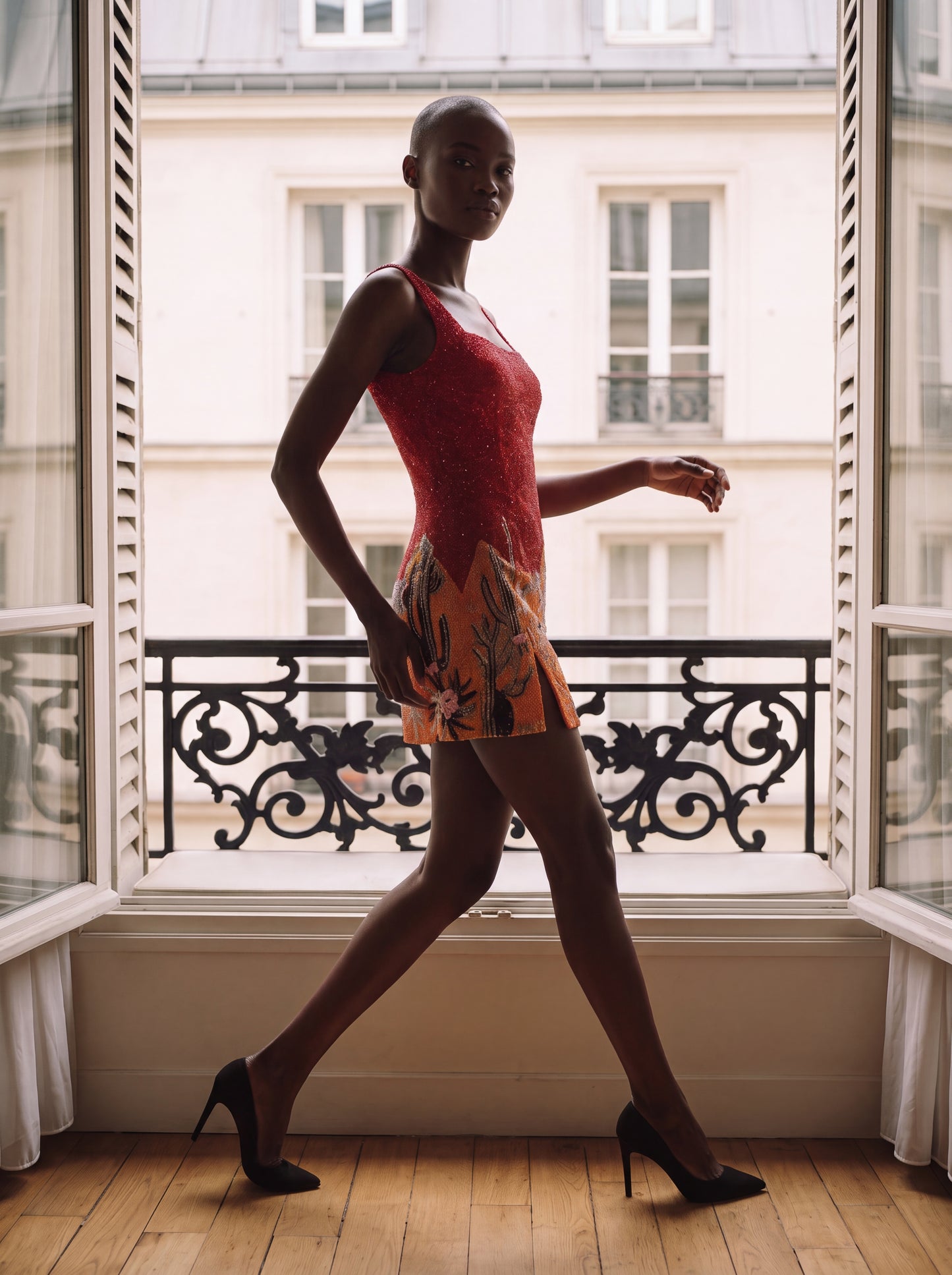 A model walks in profile by a large window wearing a sleeveless red lace mini dress with a multicolor floral and leaf print skirt, paired with black high heels, set against a Parisian-style building facade.