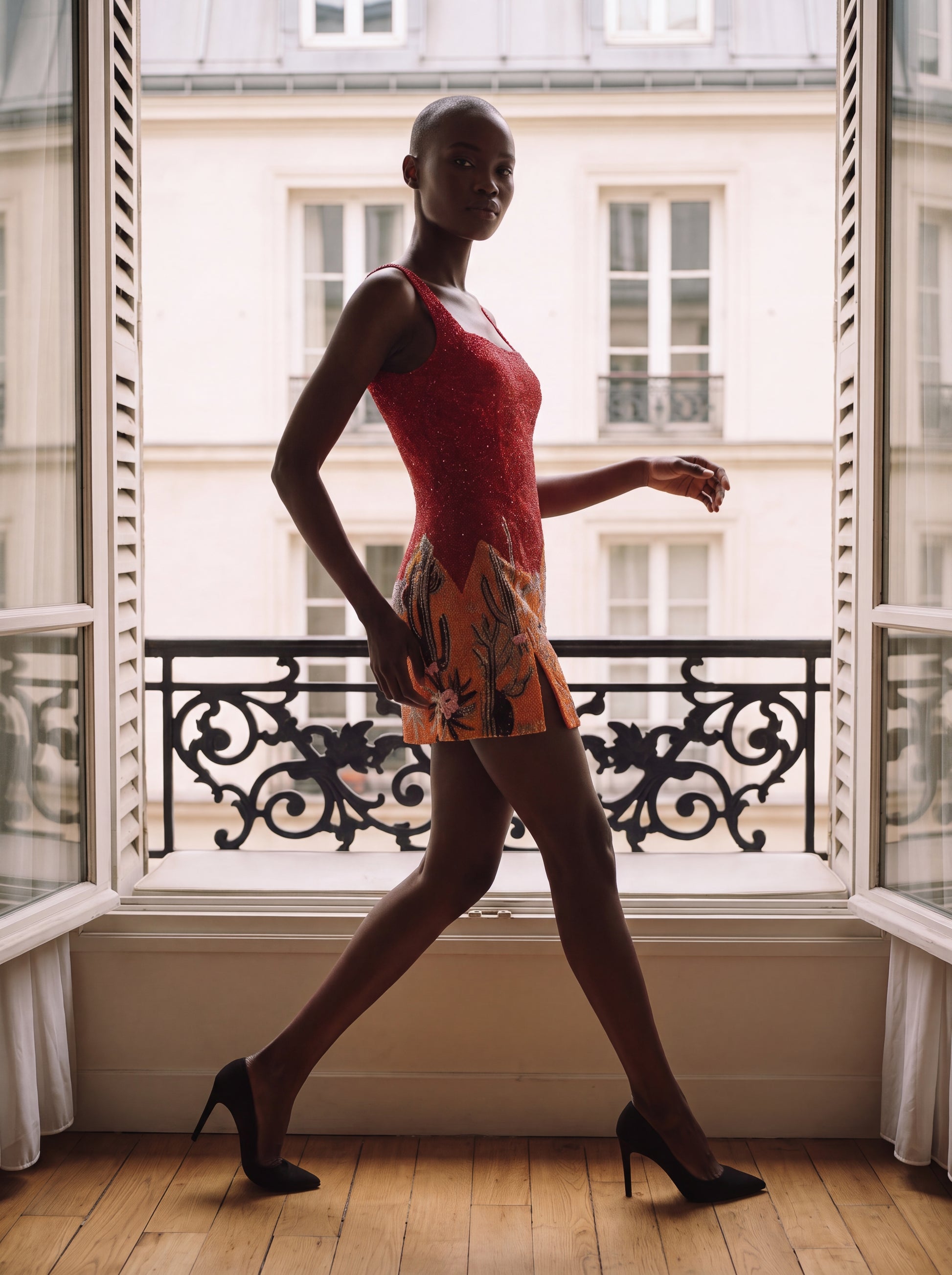 A model walks in profile by a large window wearing a sleeveless red lace mini dress with a multicolor floral and leaf print skirt, paired with black high heels, set against a Parisian-style building facade.