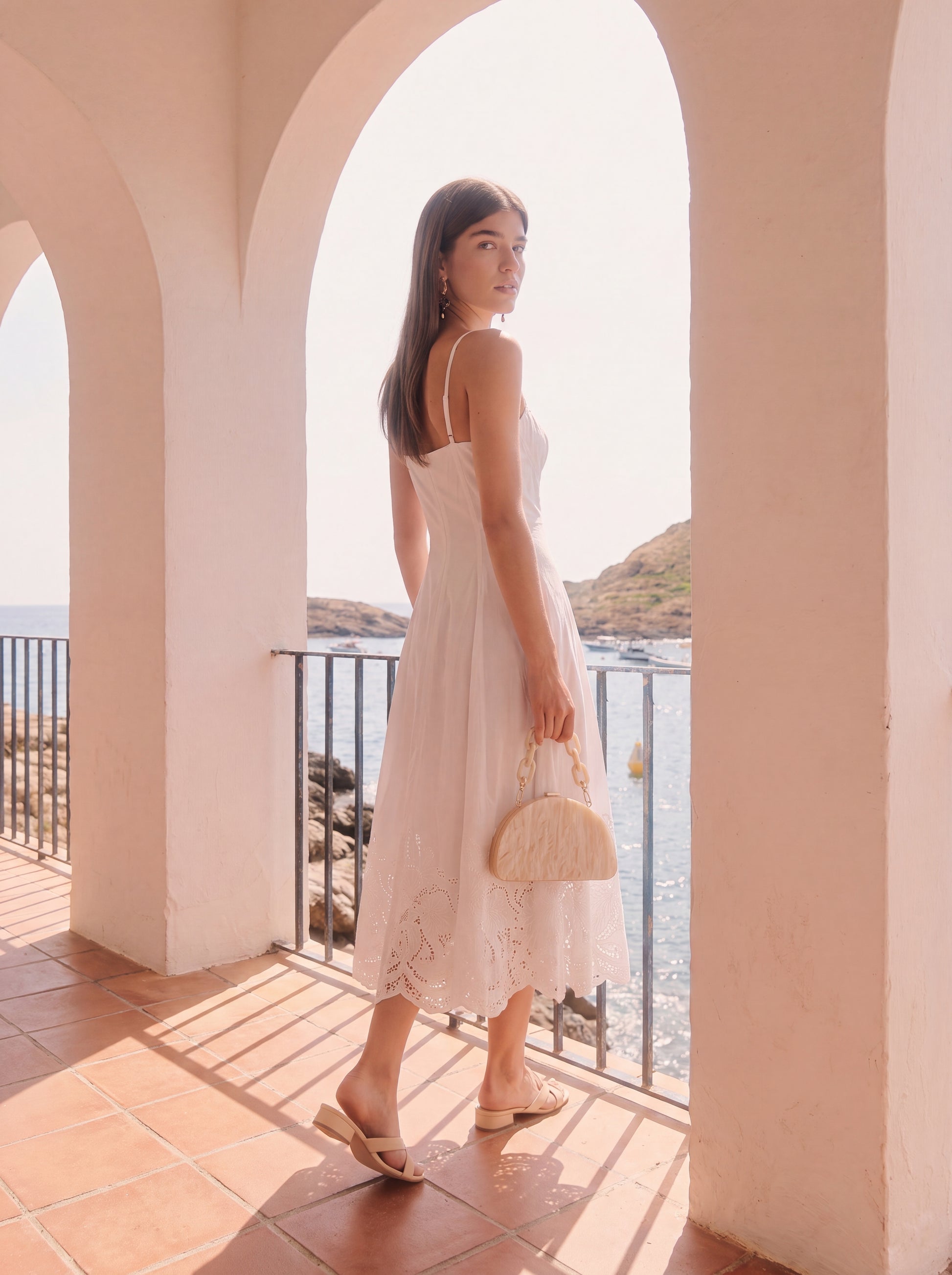 A woman wearing a light pink sleeveless midi dress stands on a sunlit terrace with arches, holding a woven straw handbag and looking over the railing toward the sea.