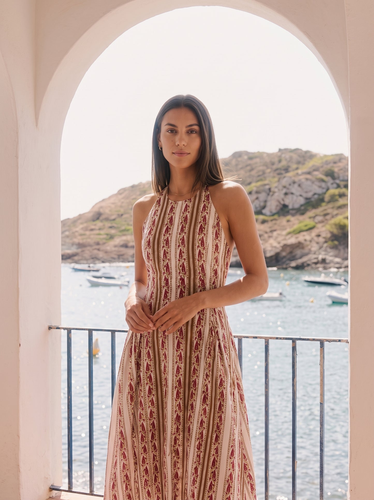 A woman wearing a sleeveless, high-neck maxi dress with a vertical red and beige pattern, standing on a balcony with a scenic coastal view of water, boats, and hills in the background.