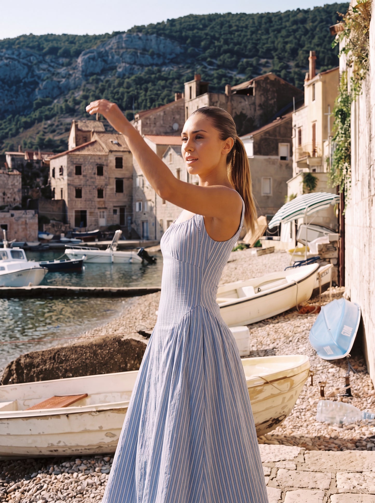 A woman wearing a light blue and white vertically striped sleeveless maxi dress stands on a stone pier in a coastal village setting with boats and buildings in the background.