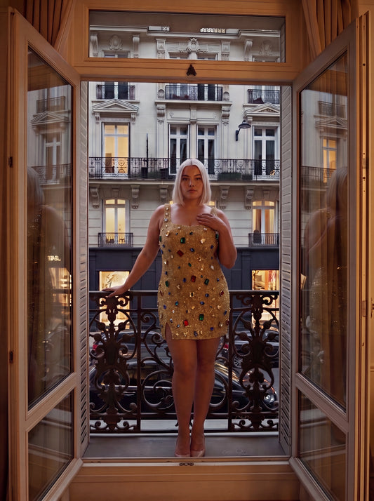A woman stands on a balcony wearing a short, embellished gold mini dress with colorful sequins, framed by open wooden doors looking out onto an urban street scene.