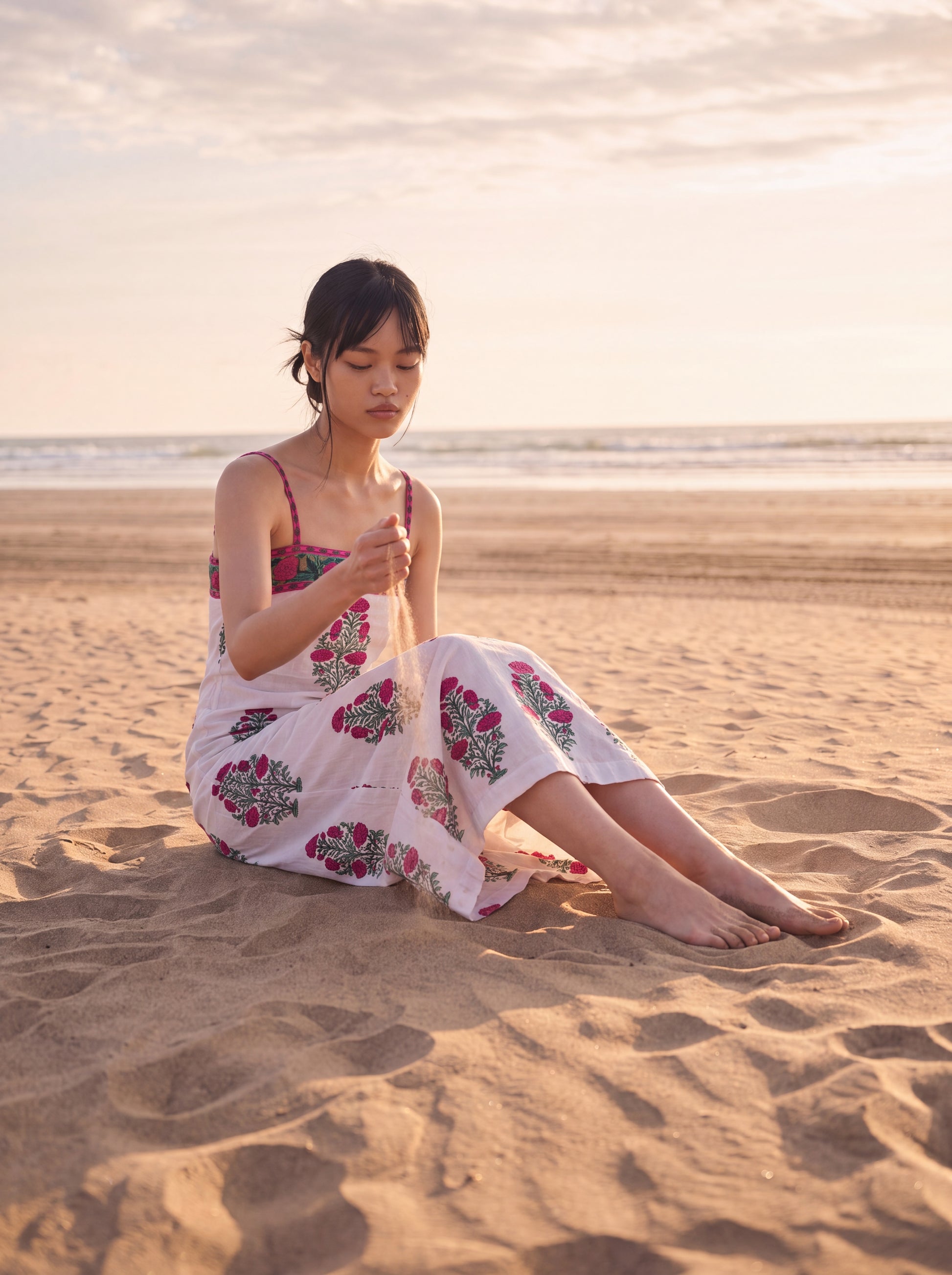 A woman sits on a sandy beach during golden hour, wearing a white sleeveless maxi dress with a colorful floral pattern. She is looking down at sand in her hands, with the ocean and cloudy sky in the background.