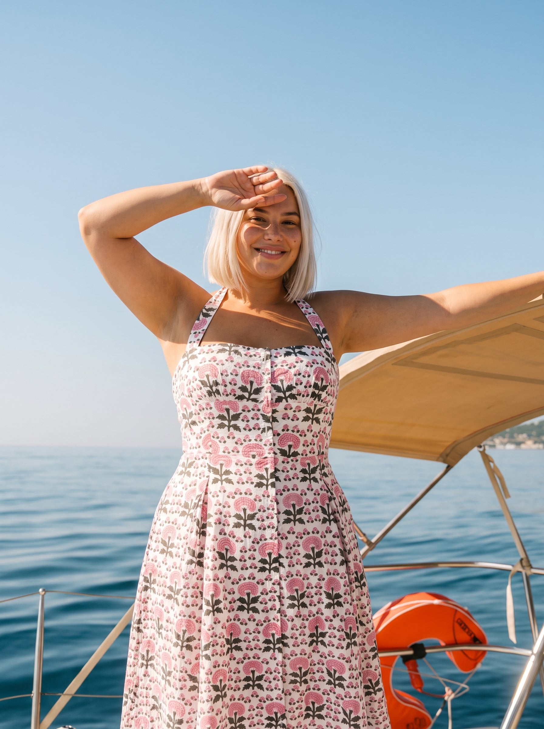 Woman wearing a pink floral halter midi dress with a fitted bodice, flowing skirt, and vintage-inspired floral print on a sailboat by the ocean.