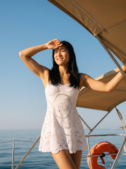 A woman wearing a white eyelet lace mini dress stands on a boat under a beige canopy, looking up and smiling with her hand over her sun. The background features a clear blue sky and sea.