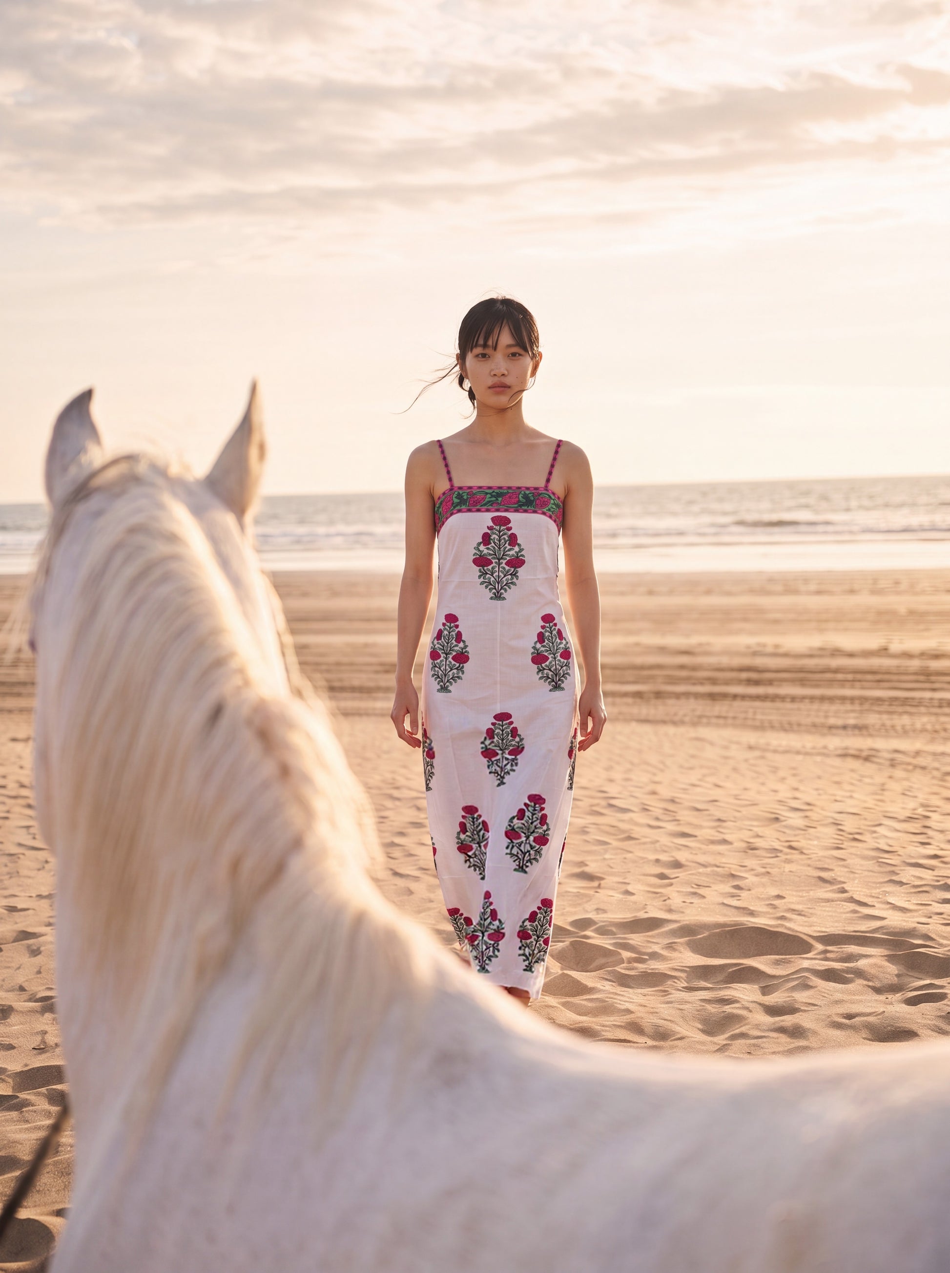 A woman stands on a sandy beach wearing a white maxi dress with colorful floral embroidery, with a large white horse in the foreground and the ocean under a warm sunset sky.